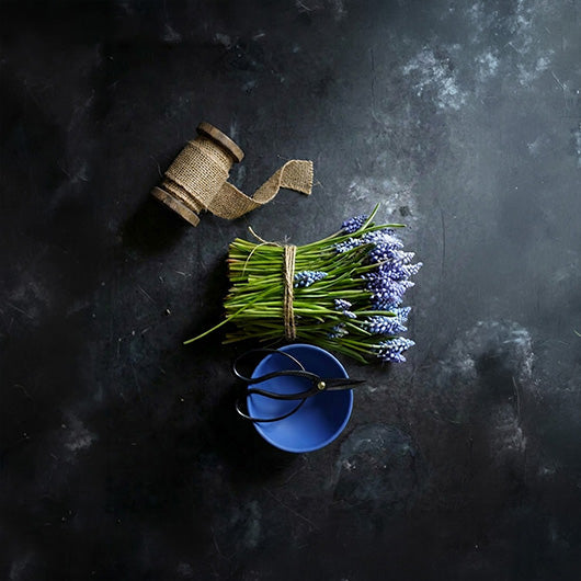A styled flat-lay of a small bouquet of purple grape hyacinth flowers tied with twine, a spool of burlap ribbon, black-handled scissors, and a small blue Baburu bowl on a dark textured background.
