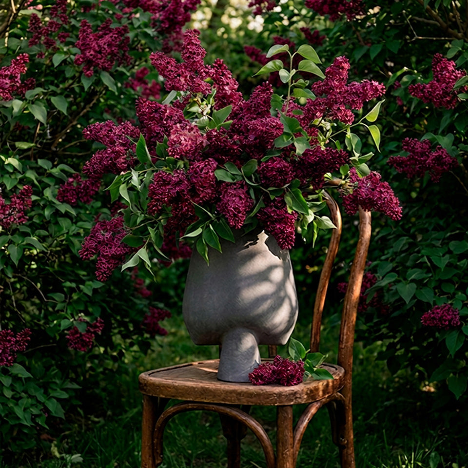 A gray Sphere sculptural vase filled with dark purple lilac stems and green leaves, placed on a rustic wooden chair in a garden setting with blooming lilac bushes.