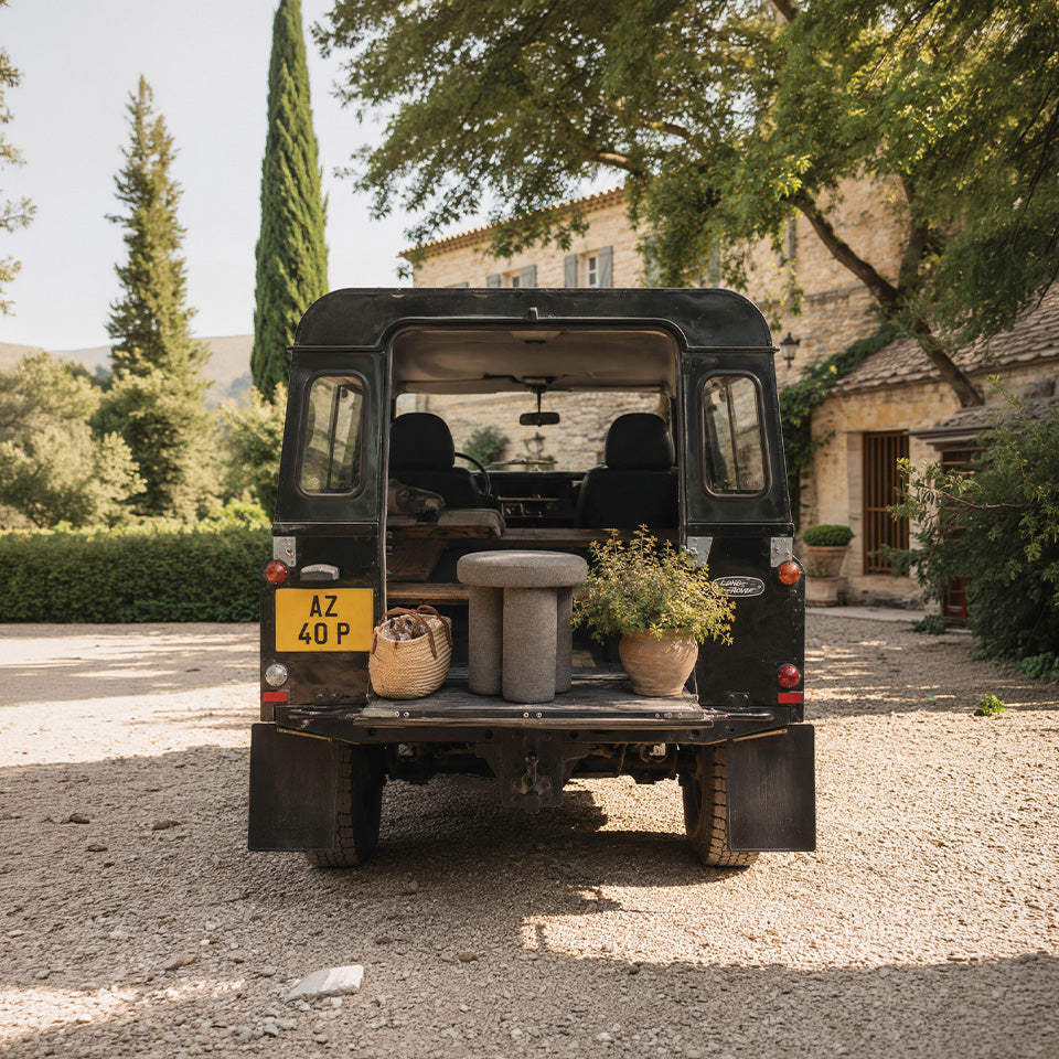 A black Land Rover utility truck parked outdoors at a stone villa, with its rear doors open to display a rustic interior setup including a gray Big Foot stool, a woven basket, and potted plants.