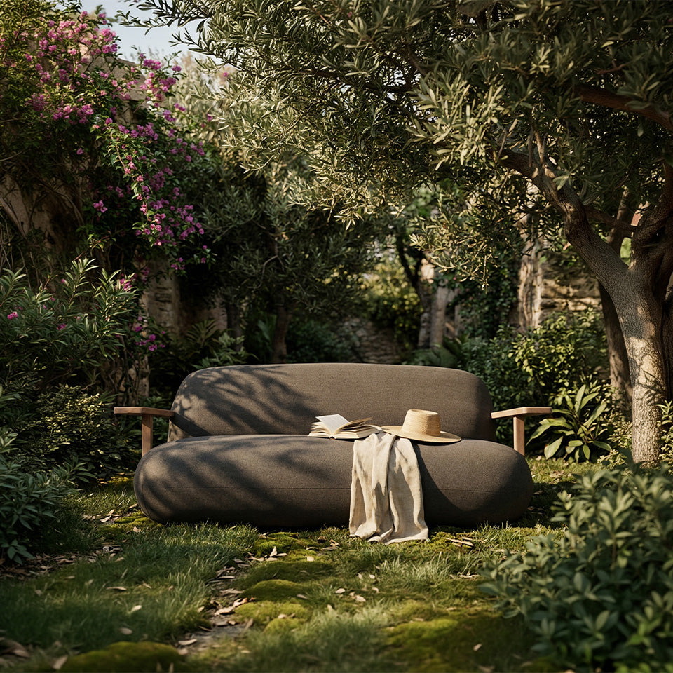 A dark grey Toe sofa outdoor lounge sofa in a lush garden setting, styled with a beige throw blanket and a straw hat.