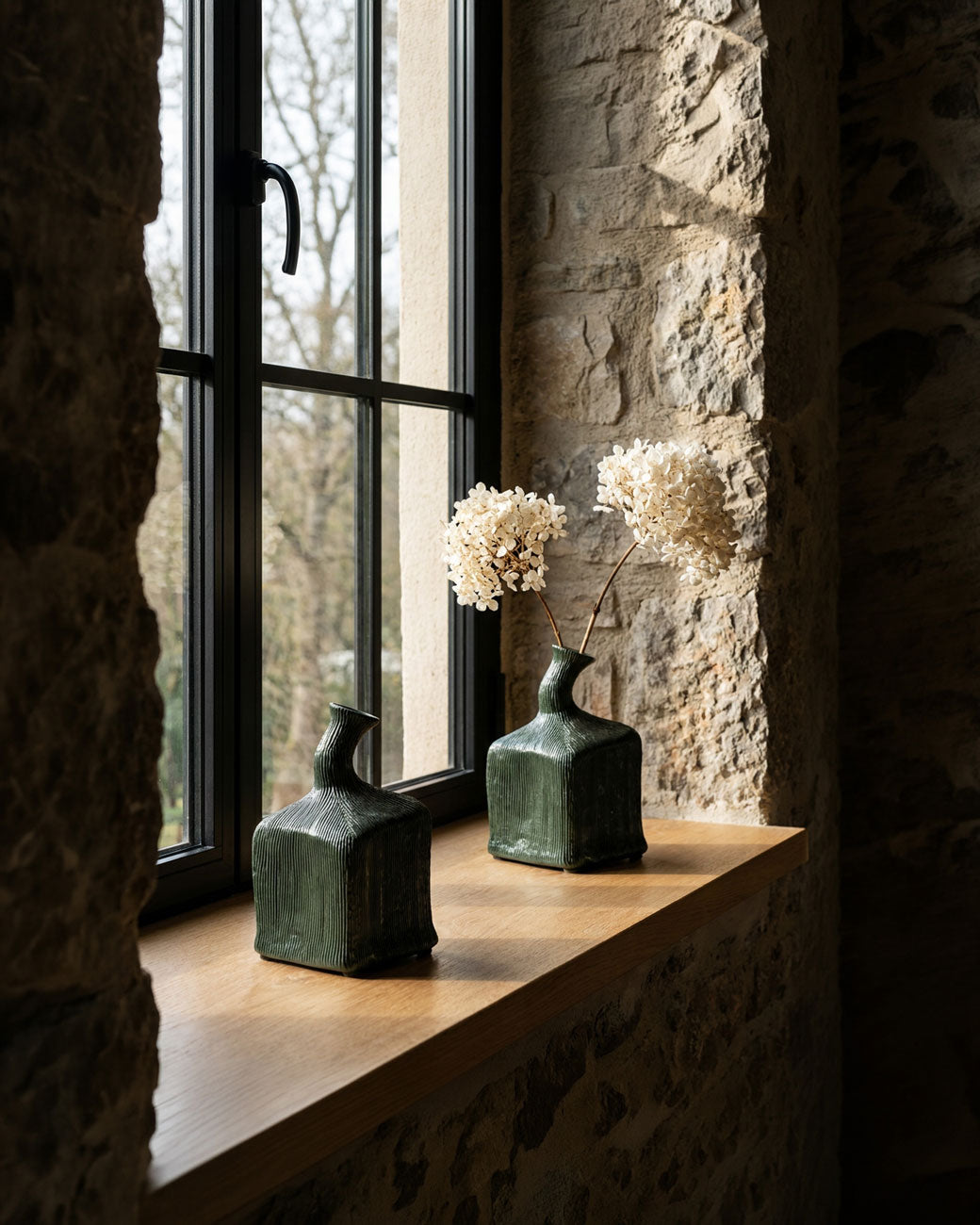 Two dark green textured ceramic Kubi vases with dried white flowers in one, displayed on a wooden windowsill in a rustic stone room.