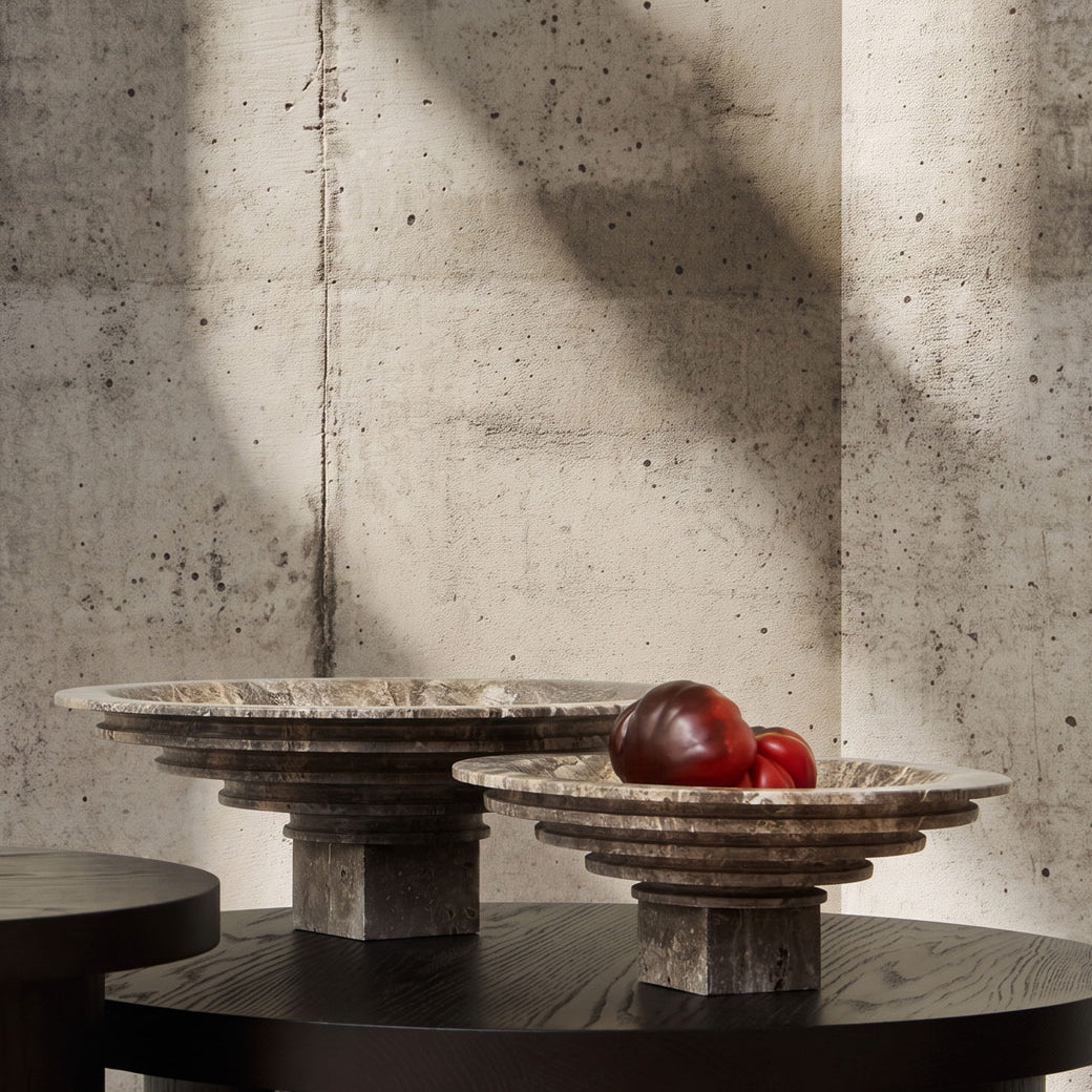Two round Sori Trays in Silver travertine with tiered rims sit on a dark wooden table against a textured concrete wall, with one bowl holding two red tomatoes. 