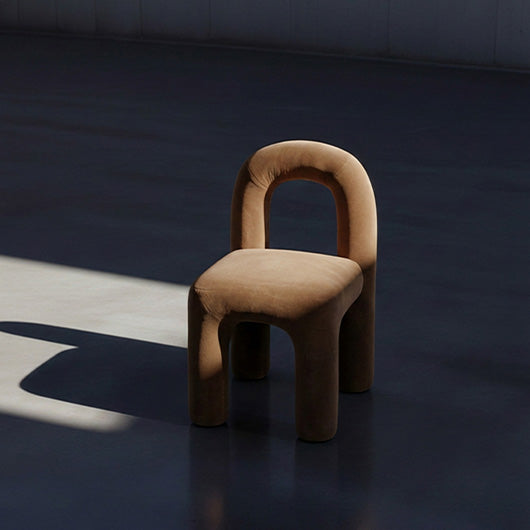 A modern upholstered Daiku dining chair with a rounded, integrated backrest and thick legs, shown in a minimalist room with dramatic side lighting and a long shadow.