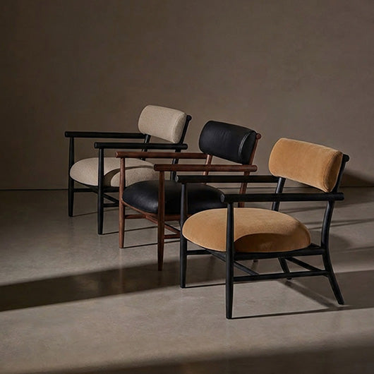Three modern Nama armchairs arranged in a row against a neutral studio backdrop. The chairs feature dark wooden frames, upholstered seats and backs in beige linen, black leather, and tan velvet, with two wooden armrests.