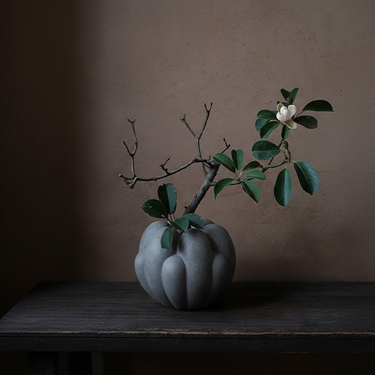 A gray Bloom ceramic vase holding a branch with green leaves and a white flower, placed on a dark wooden table against a warm brown wall.