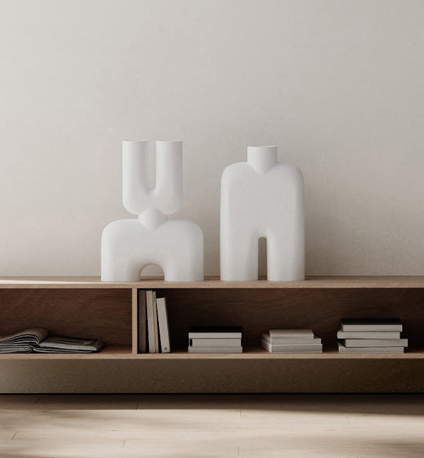 Two white abstract cobra vases on a wooden shelf against a plain wall.