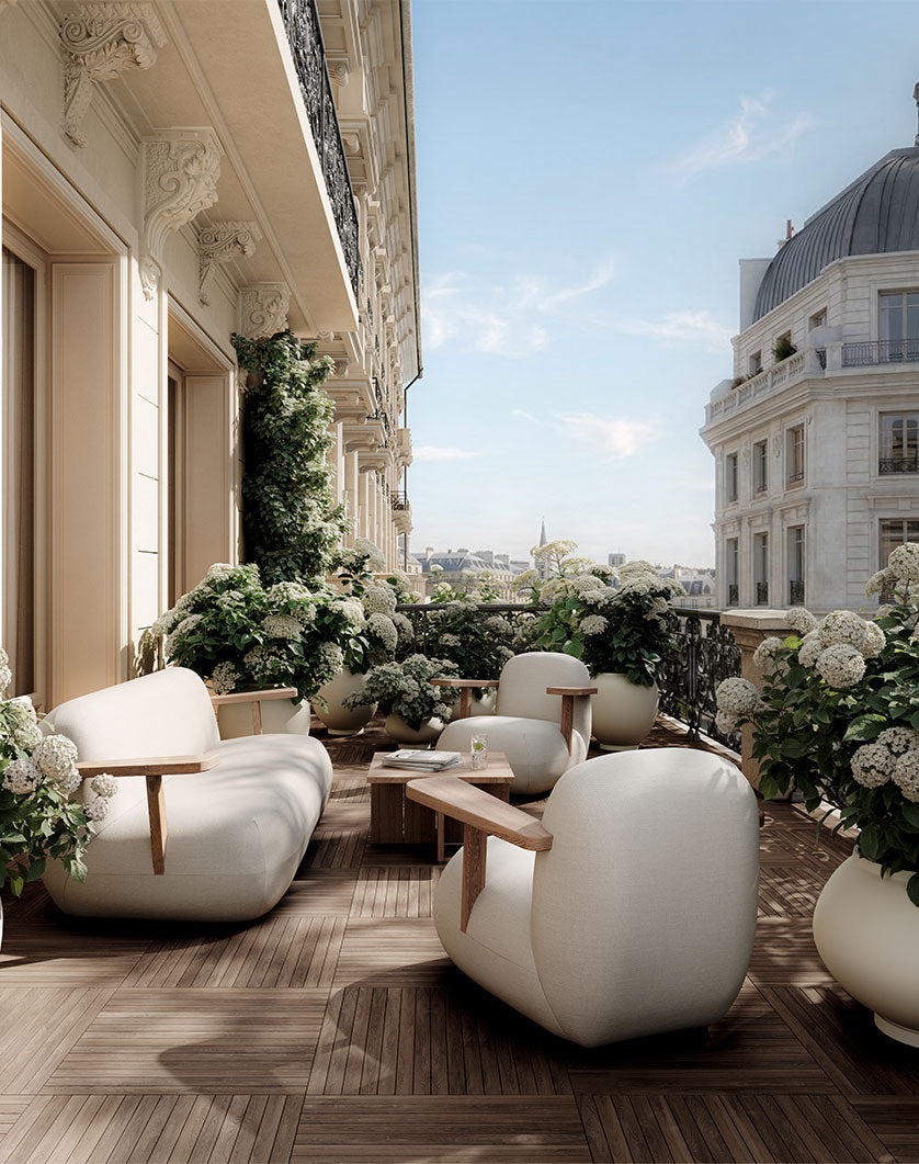 A luxury outdoor terrace furnished with modern curved light Toe lounge chairs and sofa with a small wooden Brutus side table, surrounded by Sunao Plant pots with white flowers and greenery, with a classic Parisian building facade and dome in the background under a clear blue sky.